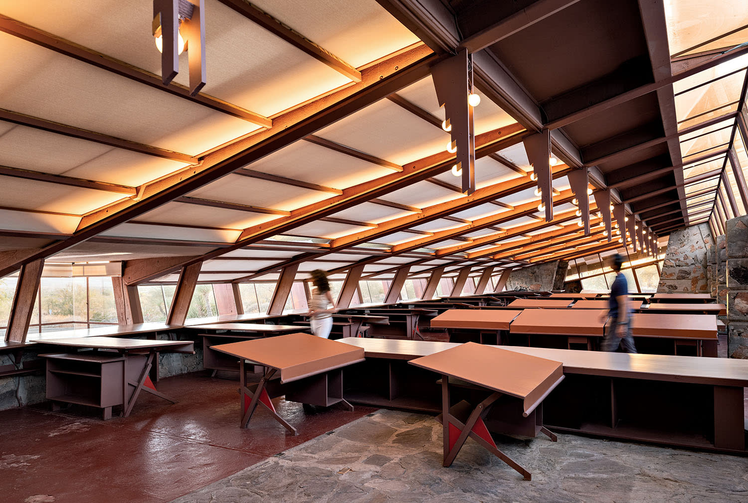 A man and woman walk through a drafting room with desks and windows.