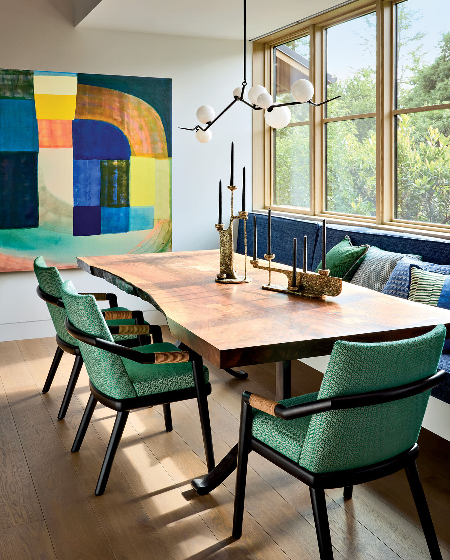 A modern dining room with a wooden table, green cushioned chairs, and a geometric abstract painting. A branch-like chandelier hangs above, adding elegance.
