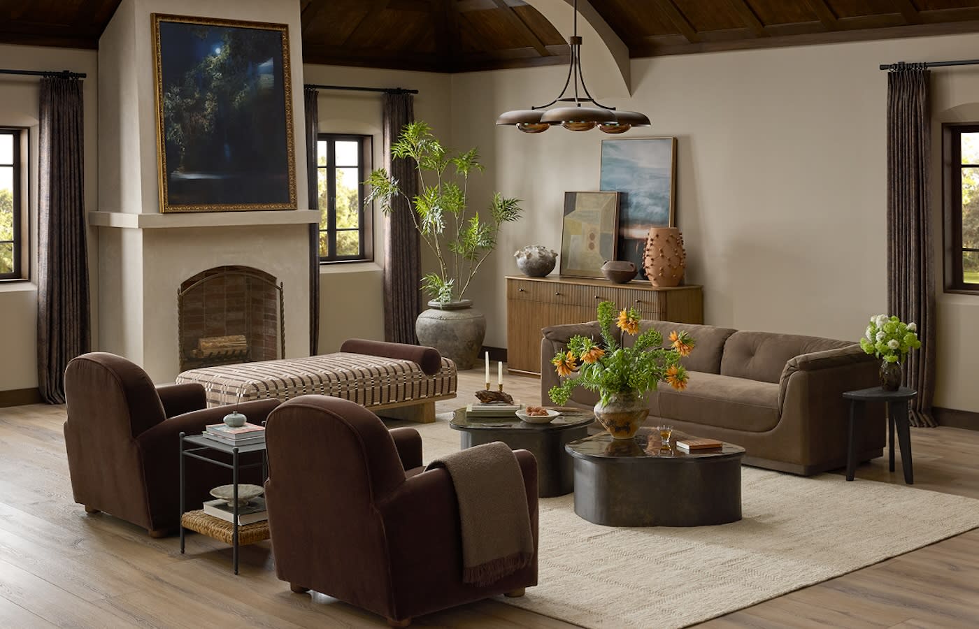 A cozy living room with a rustic feel, featuring brown sofas and armchairs, a fireplace, large plants, and a wooden cabinet. Soft natural light streams in.