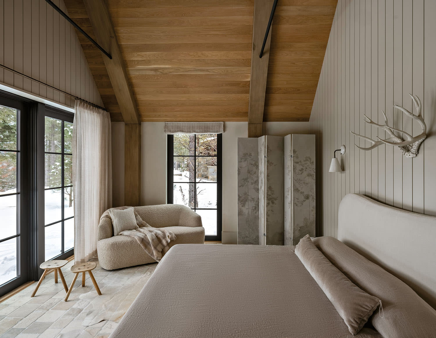 Cozy bedroom with wooden beams, soft beige tones, and a large window revealing a snowy landscape.