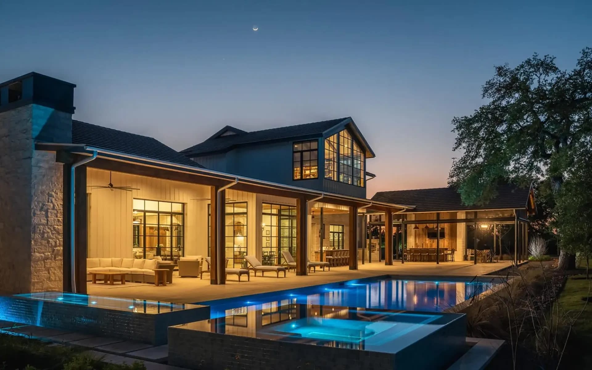 Modern luxury home at dusk with large windows glowing warmly, overlooking a sleek illuminated pool. A serene, inviting atmosphere under a crescent moon.