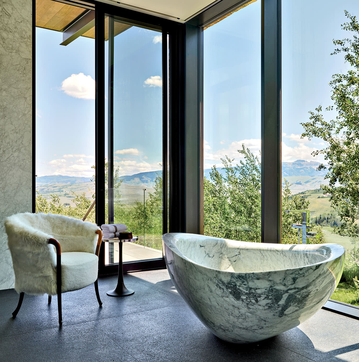 Luxurious bathroom with a marble bathtub, fur chair, and floor-to-ceiling windows overlooking a scenic landscape of trees and mountains. Serene ambiance.