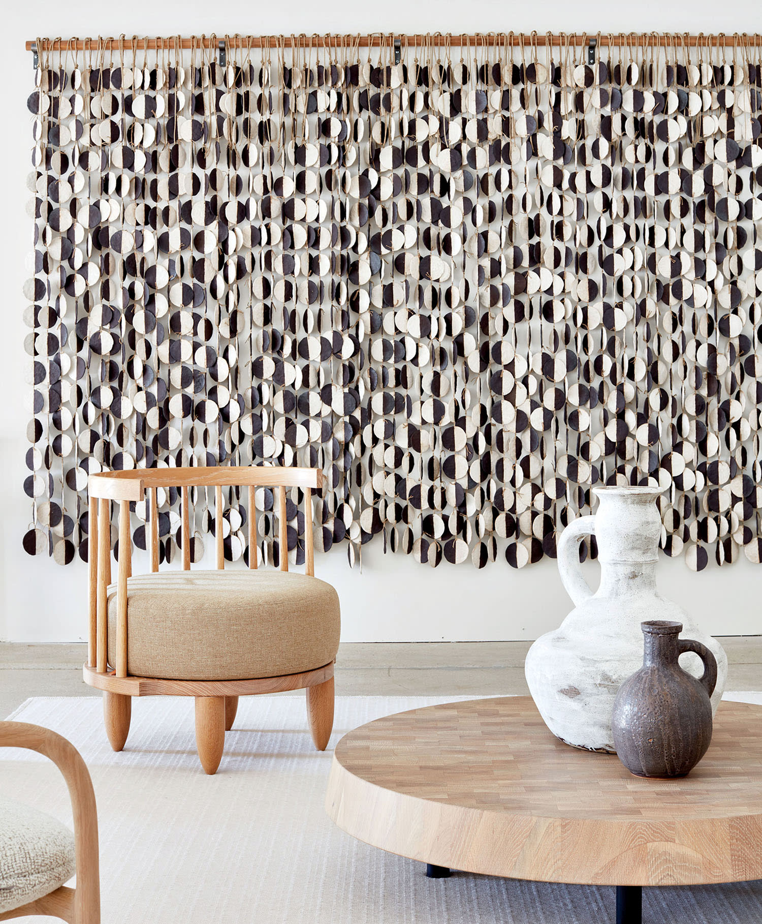 White and black vases sit on a wooden low table near chairs and a decorative wall hanging.