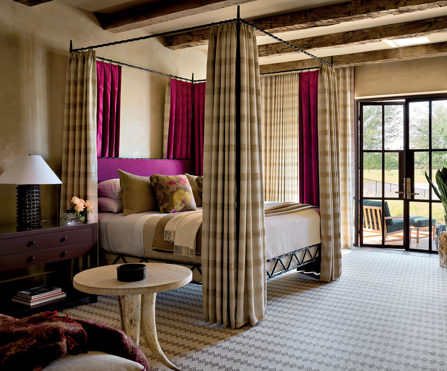 Cozy bedroom with a four-poster bed draped in beige and purple curtains. A plush chair, wooden beams, and a sunlit patio add a warm, inviting feel.