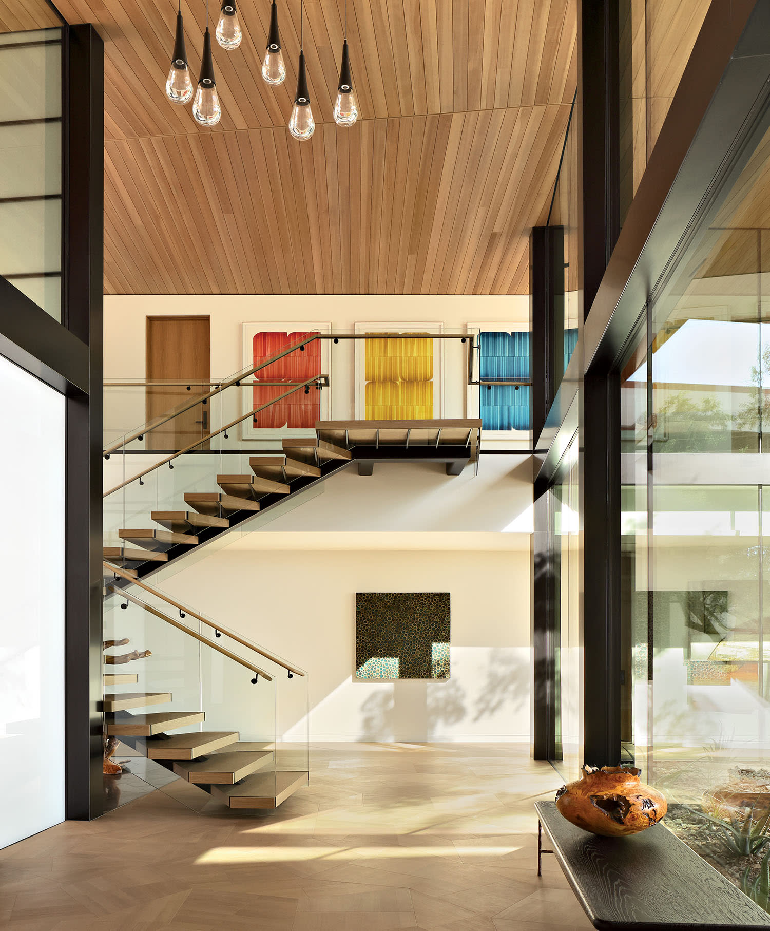 Modern interior with a wooden ceiling, spiral staircase, and large windows. Colorful wall art and an elegant bench with a vase create a warm, inviting atmosphere.