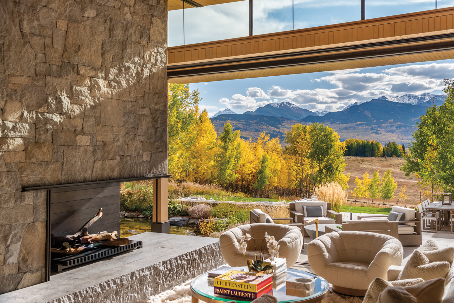 Two white, round armchairs made for relaxing and a glass cocktail table next to the granite fireplace, with open window wall behind with panoramic mountain views