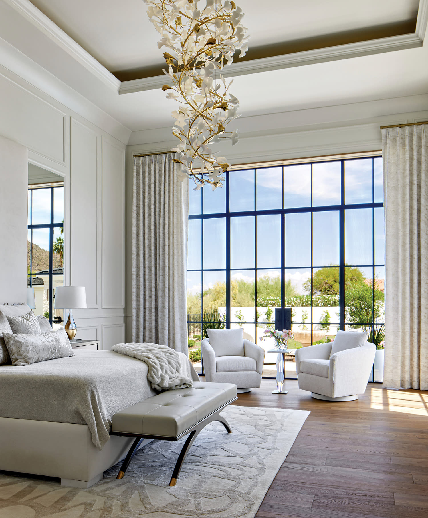 Elegant bedroom with a large window, featuring a neutral palette. A plush bed, cozy armchairs, and a modern chandelier convey a serene, luxurious ambiance.