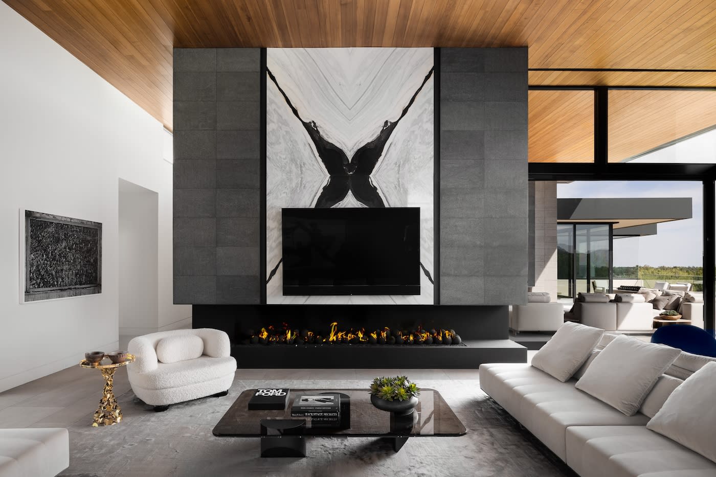 Modern living room with a sleek black TV mounted on a geometric stone wall above a linear fireplace. White sofas and armchairs sit on a neutral rug, and a gold side table adds elegance. Large windows in the background provide ample natural light.