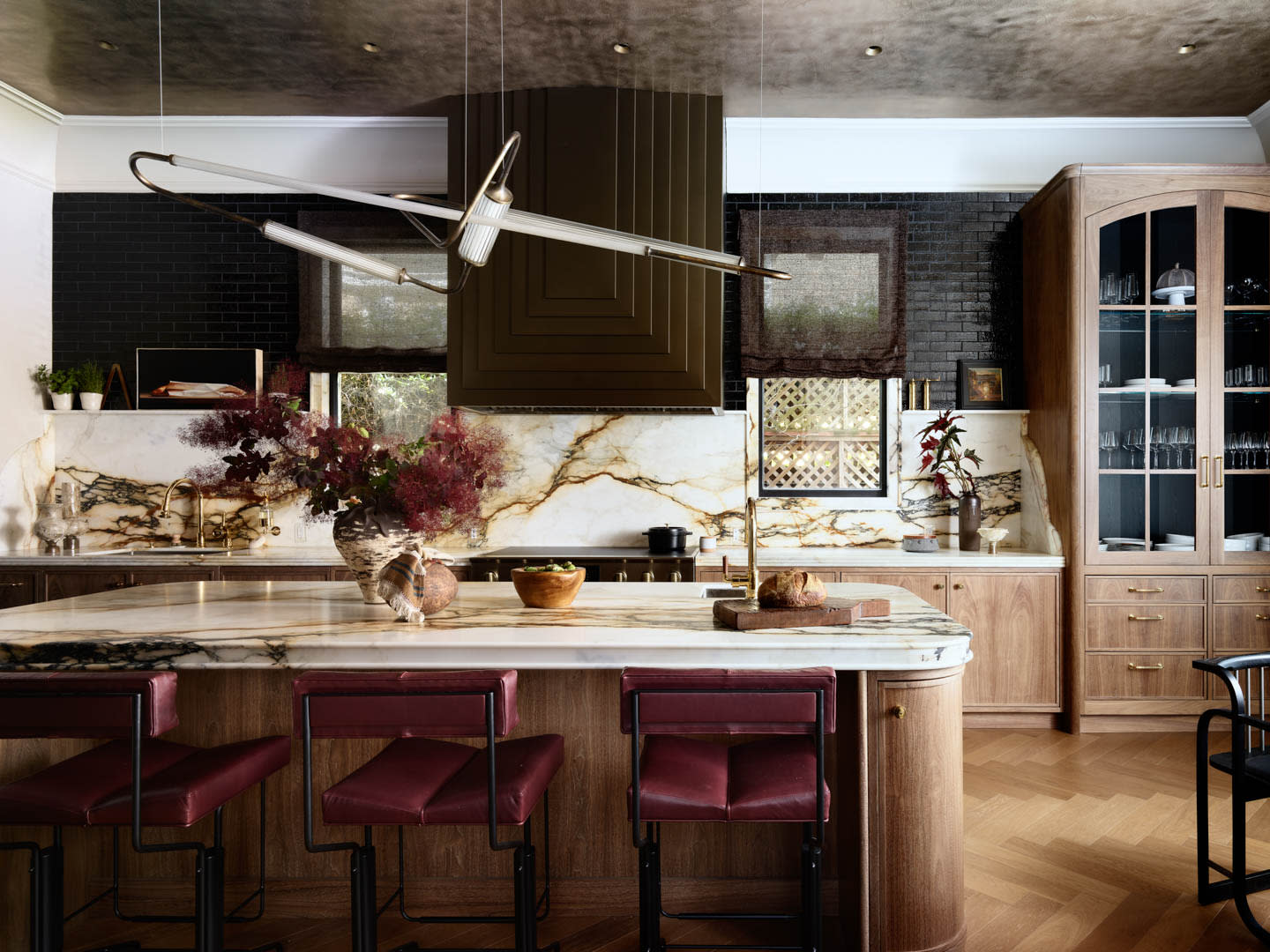 Elegant kitchen with a marble island, burgundy stools, and gold-veined backsplash. Wooden cabinets, black brick walls, and modern light fixture create a cozy yet sophisticated atmosphere.