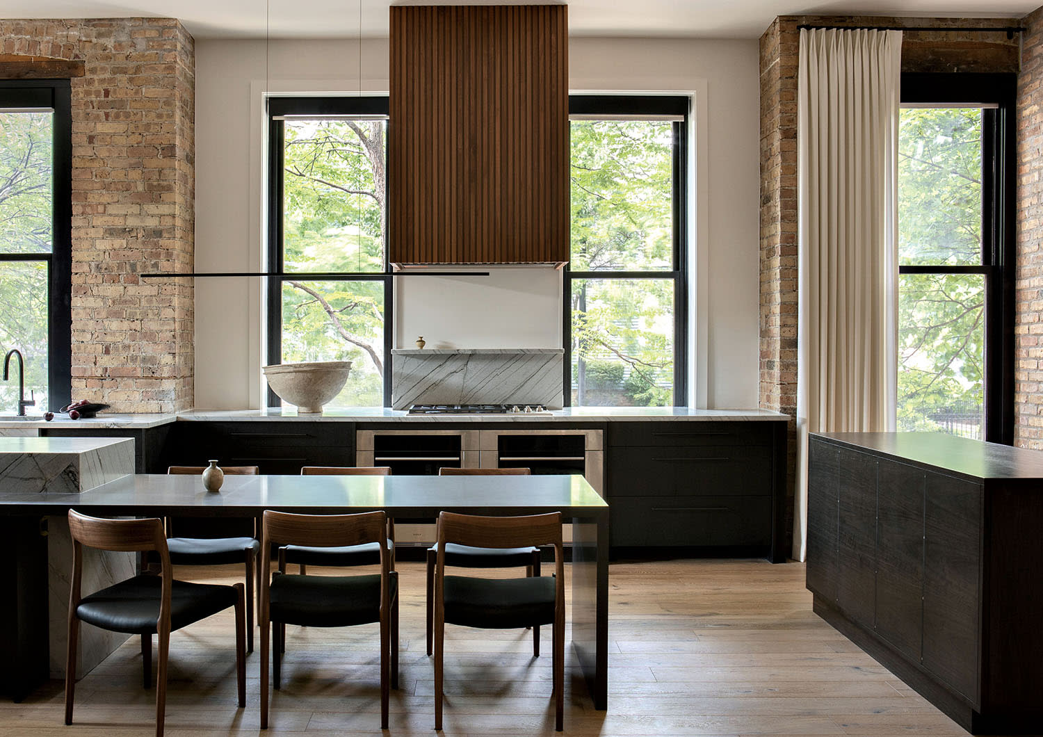 A windowed kitchen with dark cabinetry, brick walls and an adjoining dining table set with chairs.