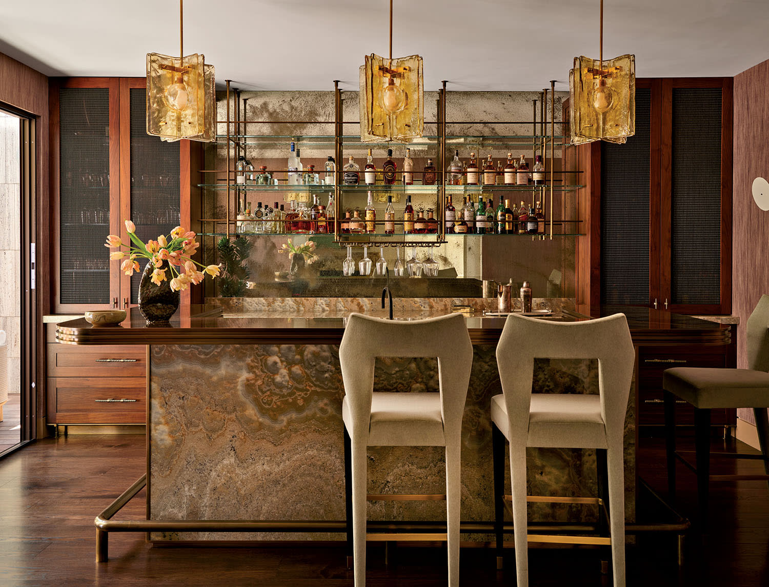Elegant home bar featuring marble countertops, plush beige stools, and a mirrored backdrop with shelves of bottles. Warm lighting from ornate chandeliers.