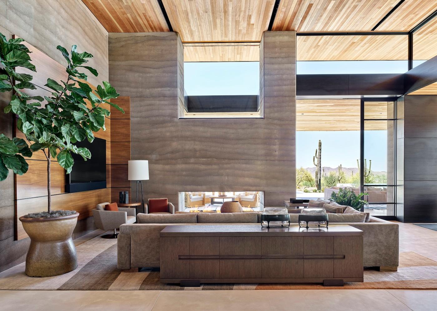Spacious modern living room with large windows, wooden ceiling, and neutral tones. Features a potted tree, plush couches, and desert views outside. Calm ambiance.