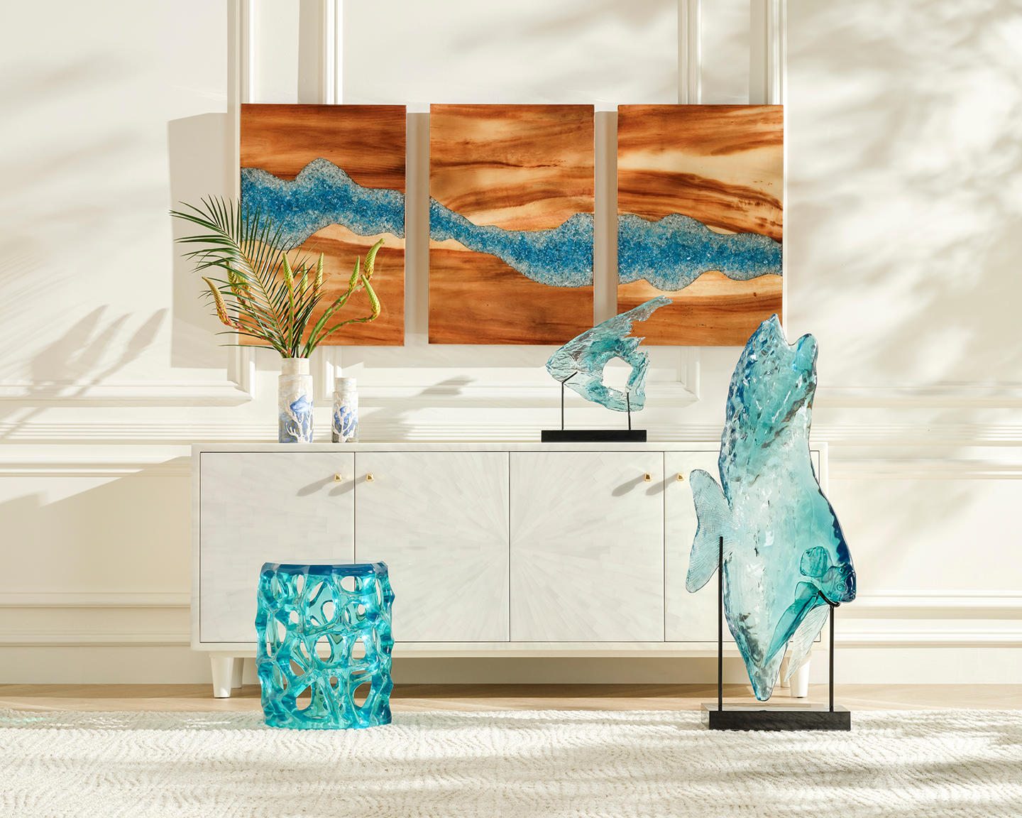 Modern room with white cabinet and vibrant blue glass sculptures. Triptych wood art with blue accents hangs above. Light casts soft shadows.