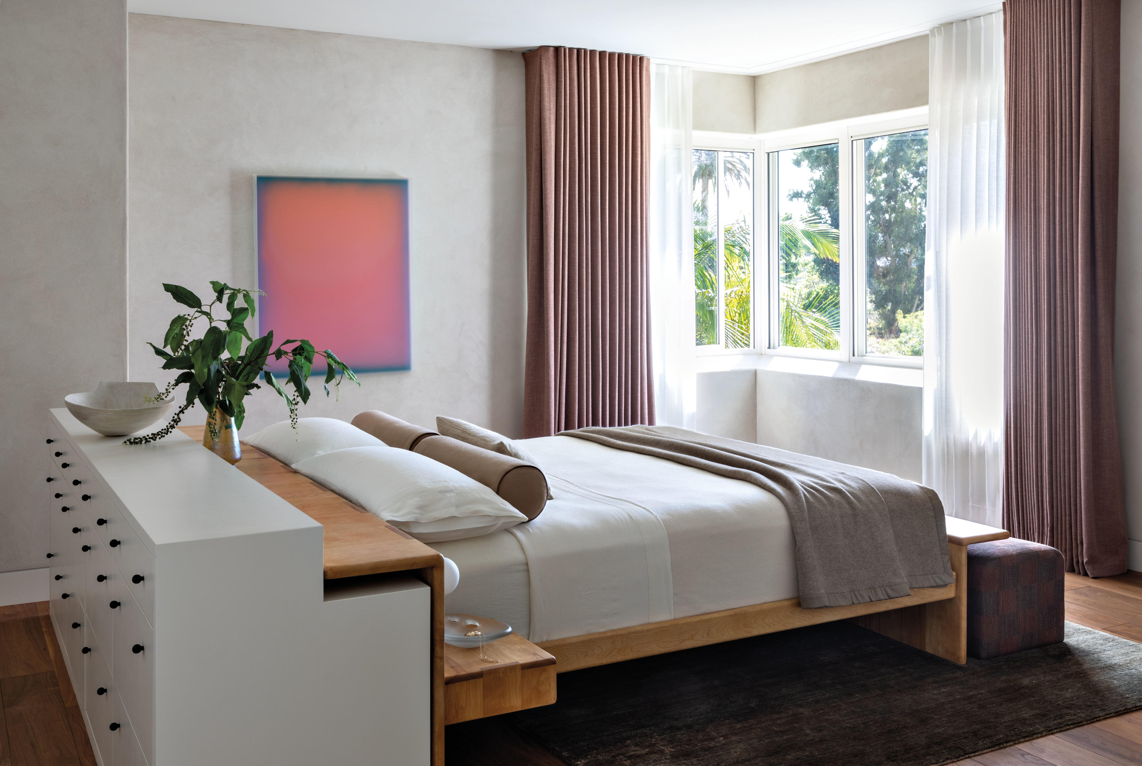 clean line bedroom with a neutral toned bed, colorful artwork and light red drapes