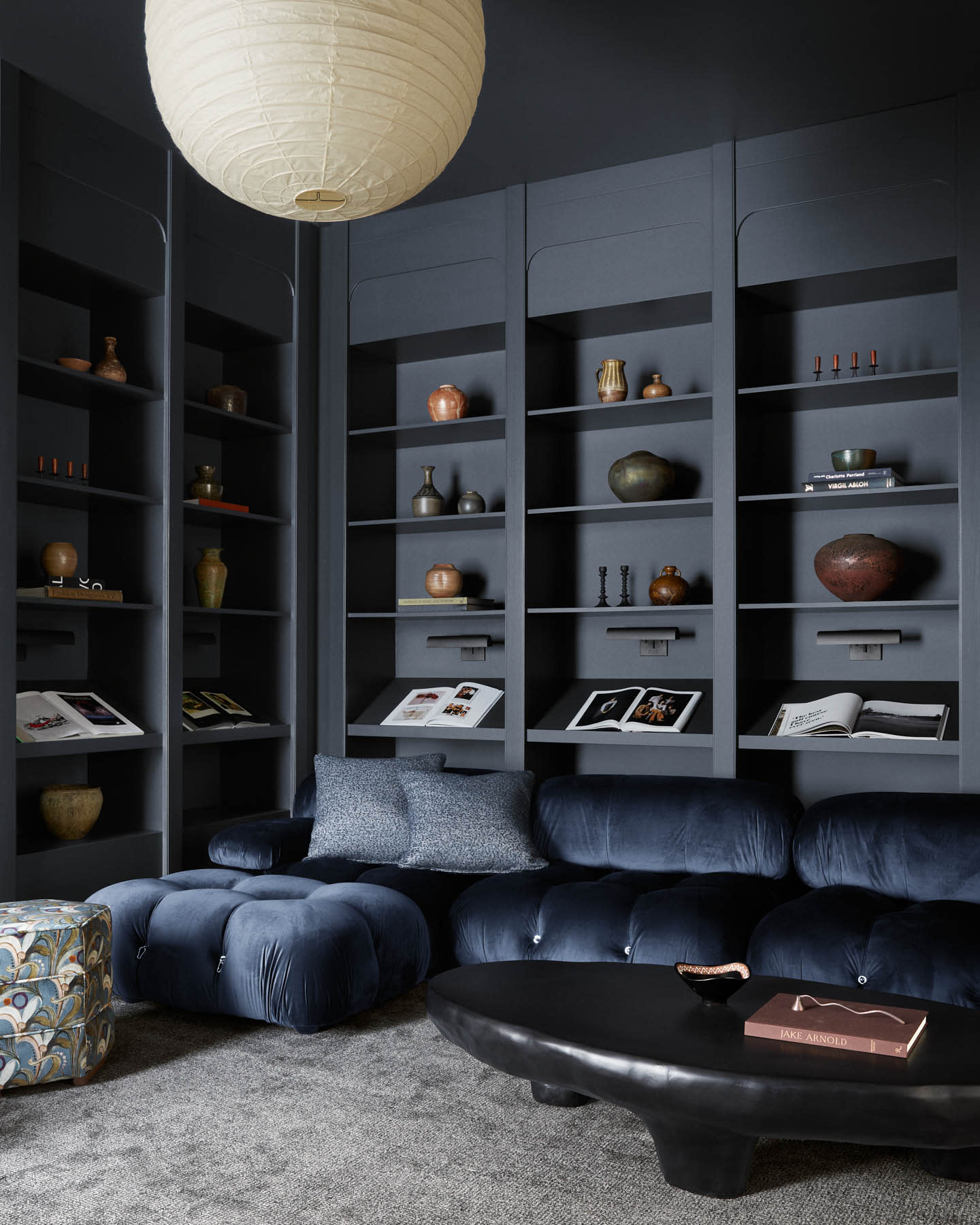 A cozy, modern living room with dark built-in shelves displaying pottery and books. A plush navy sofa and a black oval coffee table enhance the sophisticated vibe.