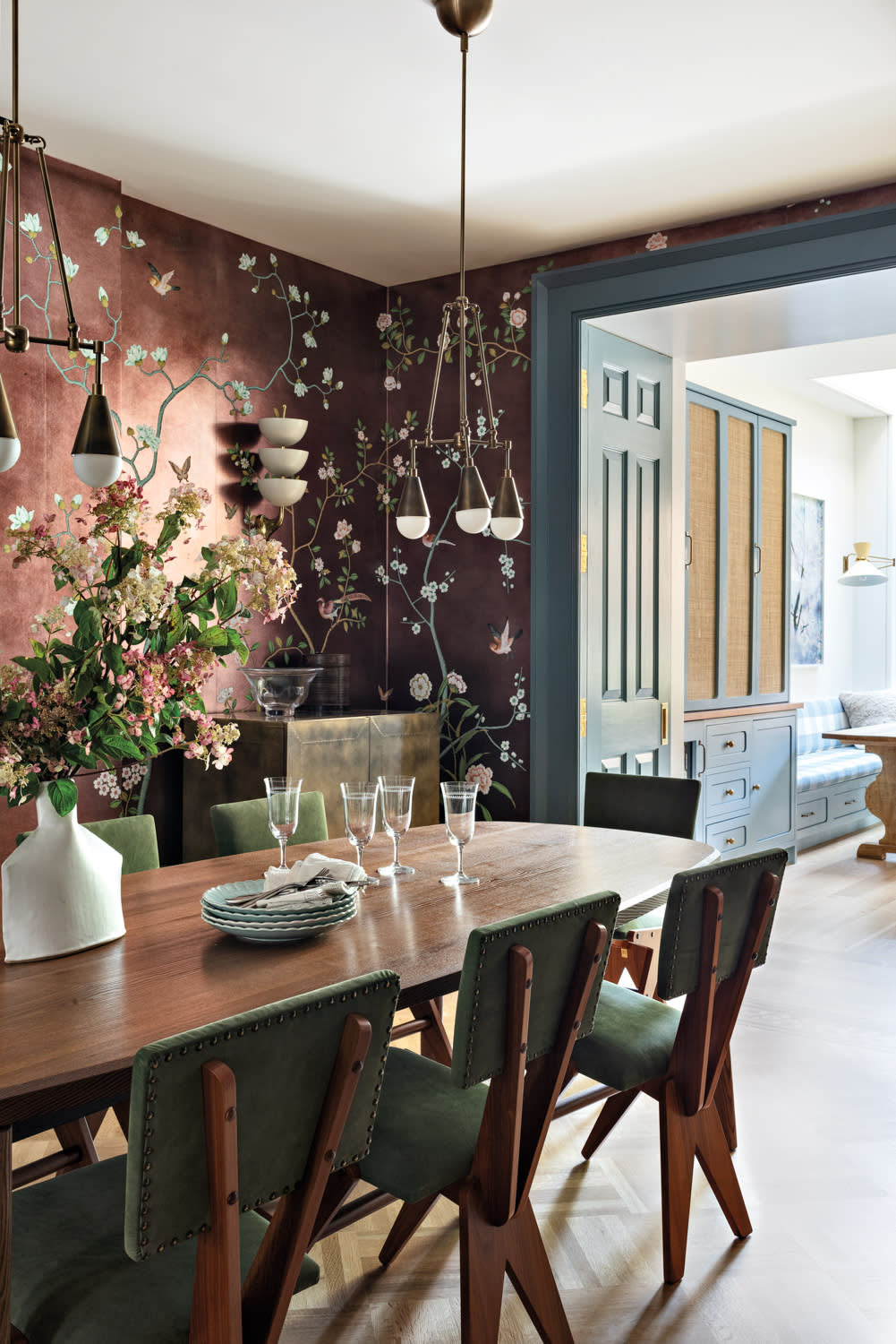 Elegant dining room with a wooden table and green chairs. Floral wallpaper creates a warm tone. Modern pendant lights hang above, creating a cozy atmosphere.
