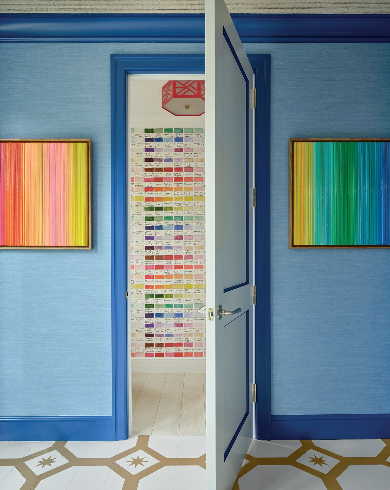 Open blue door reveals a colorful room with a vibrant paint swatch wall and geometric ceiling light. Two colorful striped paintings hang on blue walls.