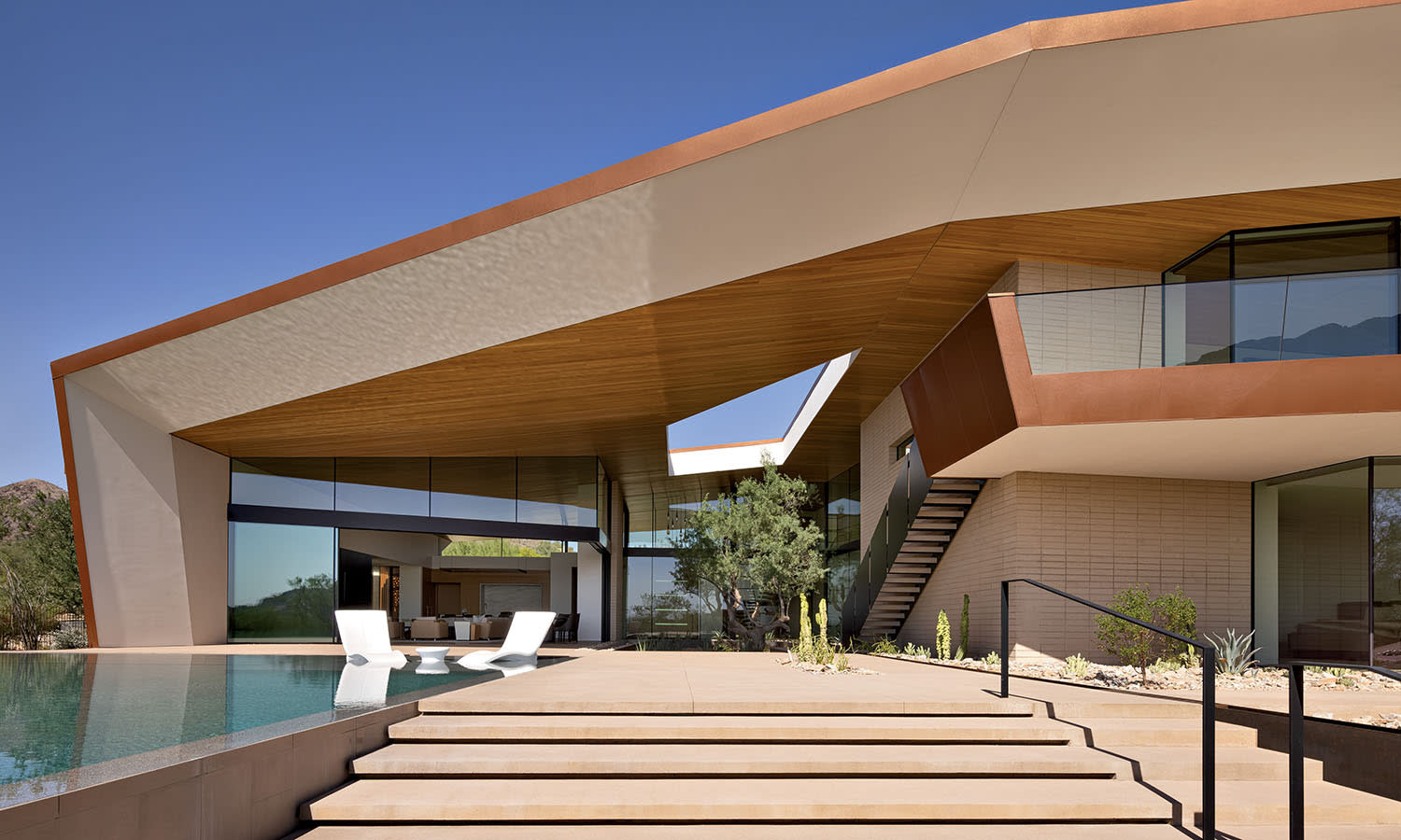 Modern angular house with an L-shaped design, featuring large glass windows, an infinity pool, and minimalist white chairs under a clear blue sky.