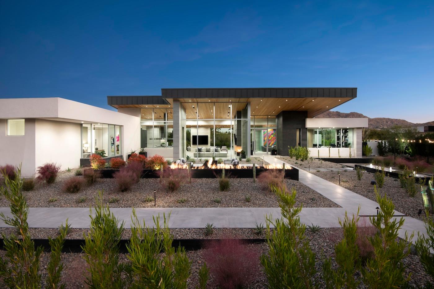 Modern house at dusk with large windows, flat roof, and sleek design. Gravel landscaping and desert plants enhance the minimalist, tranquil setting.