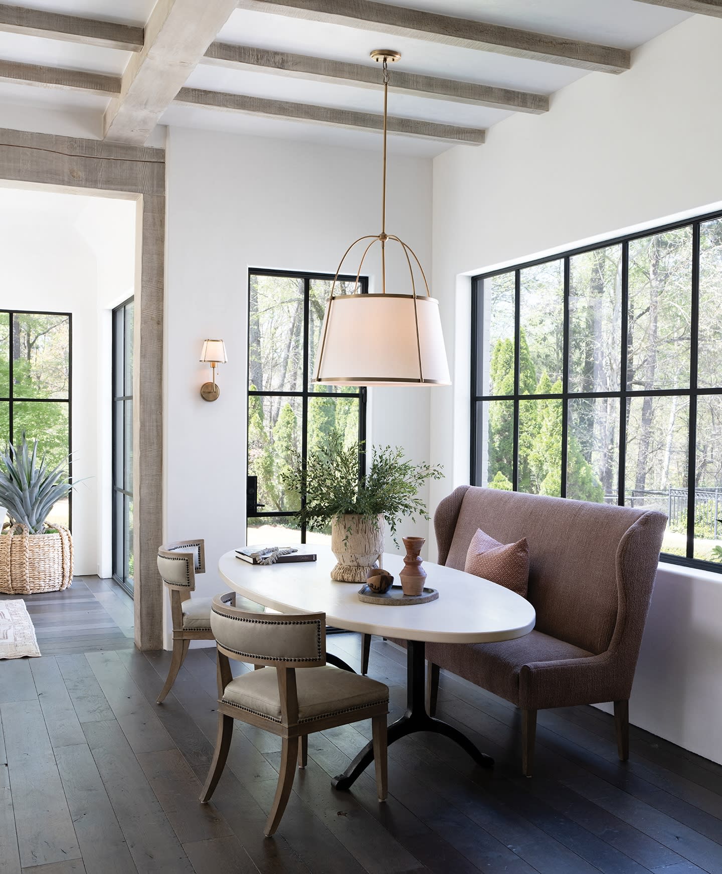 Stylish dining nook with a white oval table, two beige chairs, and a brown bench with cushions. Large windows let in natural light, and a pendant lamp hangs above.