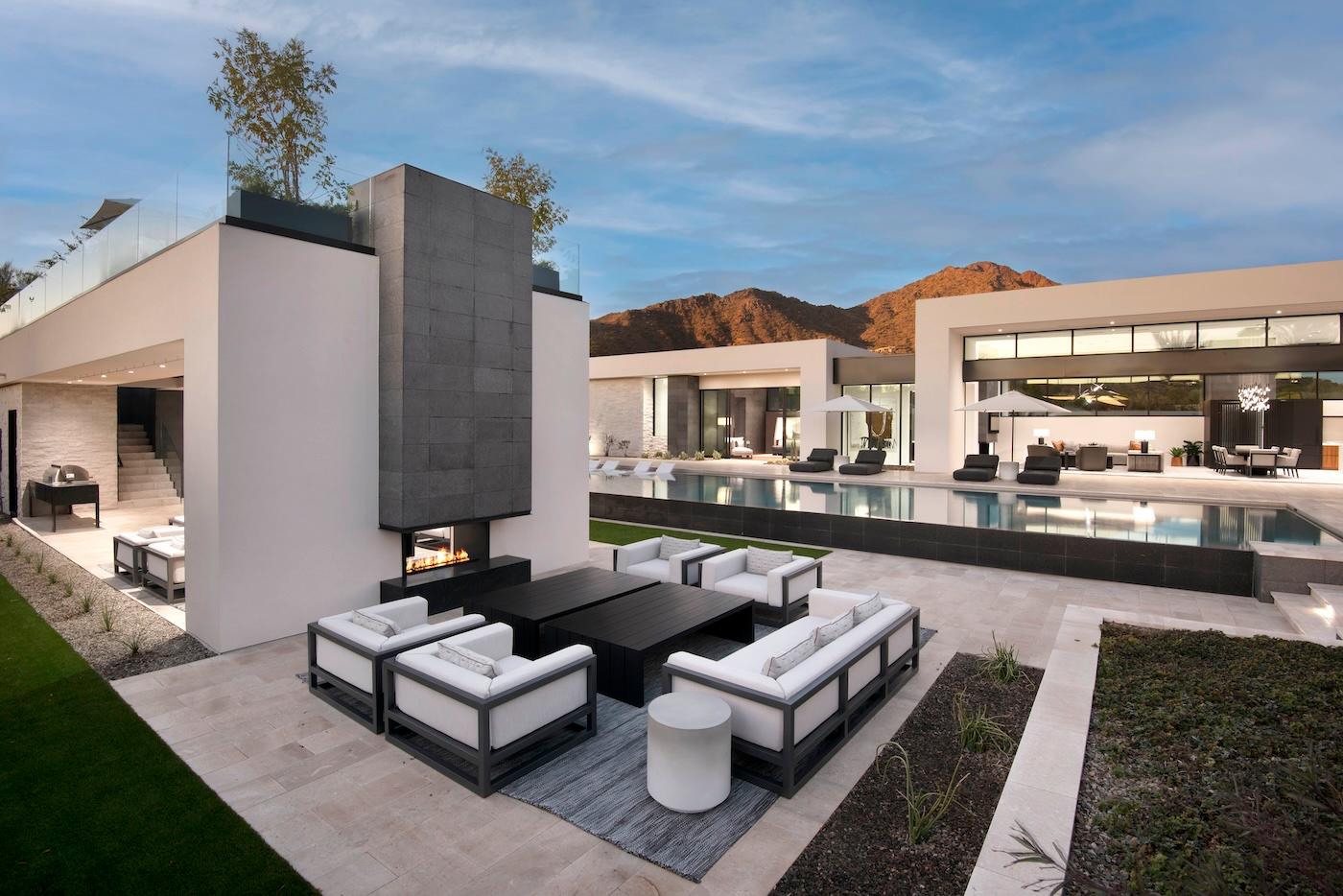 Modern luxury home with a sleek design, featuring an outdoor patio with white furniture and a fire pit. A large pool reflects the surrounding mountains.