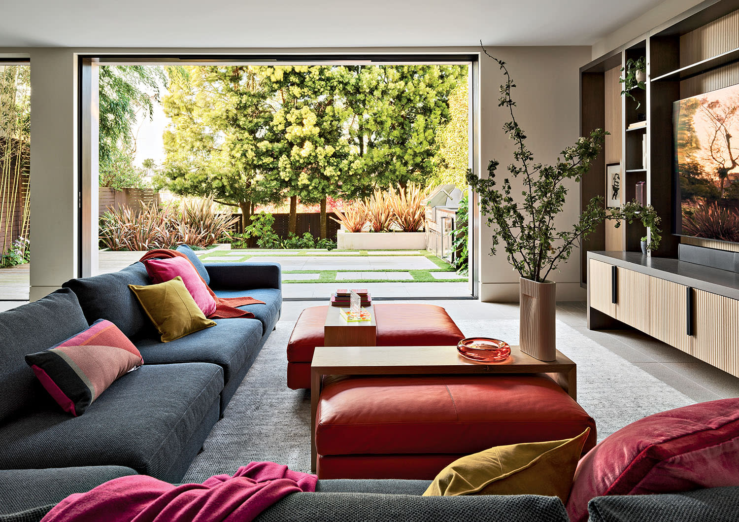 Modern living room with a large gray sectional sofa, colorful pillows, and red ottomans. Open glass doors reveal a lush green garden outside.