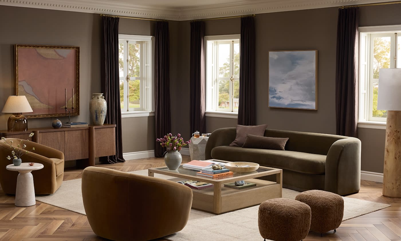 Sophisticated living room with a brown palette, featuring a velvet sofa, armchairs, and wooden coffee table. Light from large windows creates a warm, inviting atmosphere.