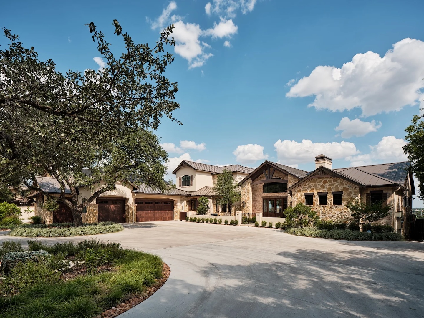 Luxurious stone house with a grand entrance, three-car garage, and lush landscaping under a bright blue sky with fluffy clouds, conveying elegance.