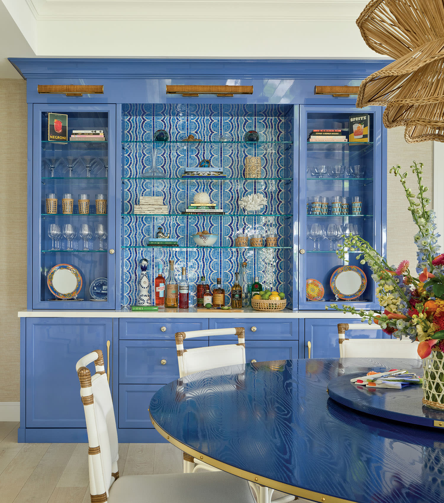 Bold blue cabinets with glass doors hold glasses and decor against patterned wallpaper. A round table with vibrant flowers adds warmth and charm.