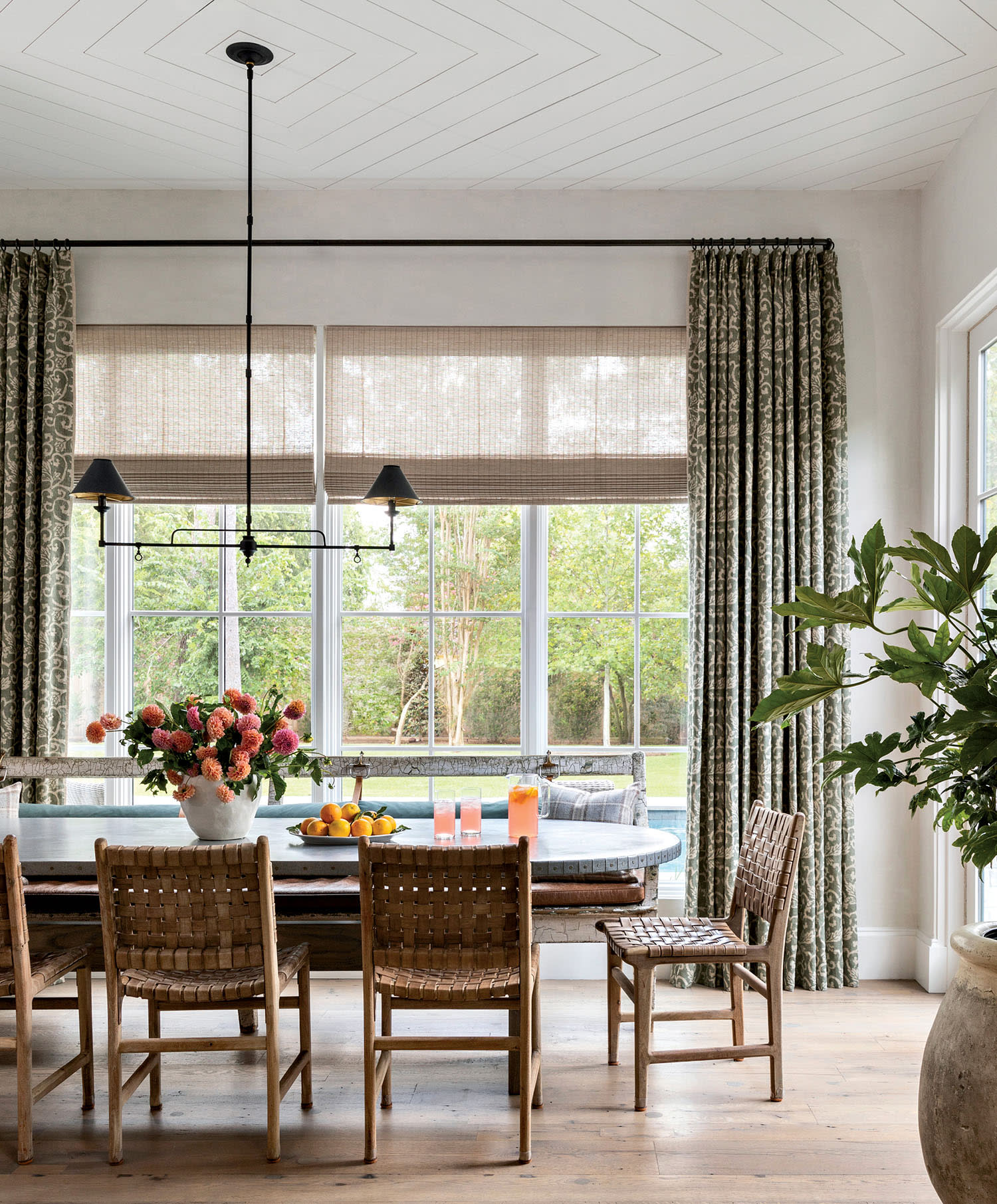 Bright dining area with woven chairs around a rustic table. Large windows reveal greenery outside.
