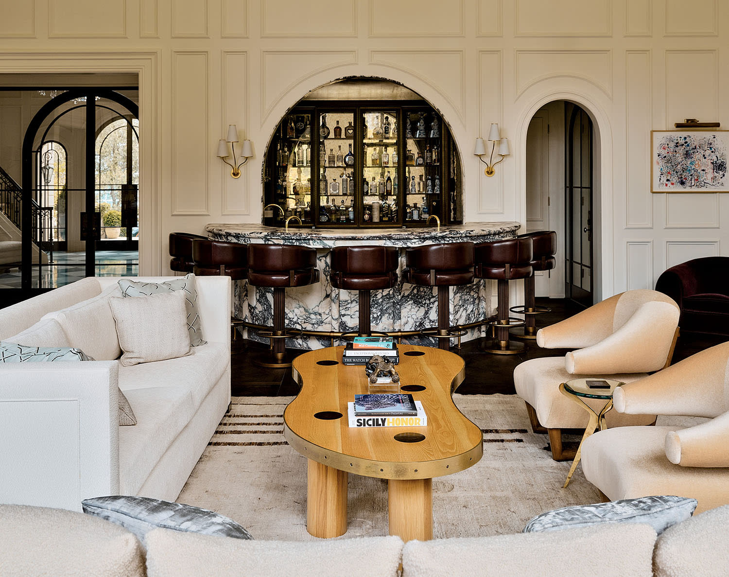 Elegant living room with a central marble bar, plush seating, a large ornate chandelier, and paneled walls. Luxurious, inviting ambiance.