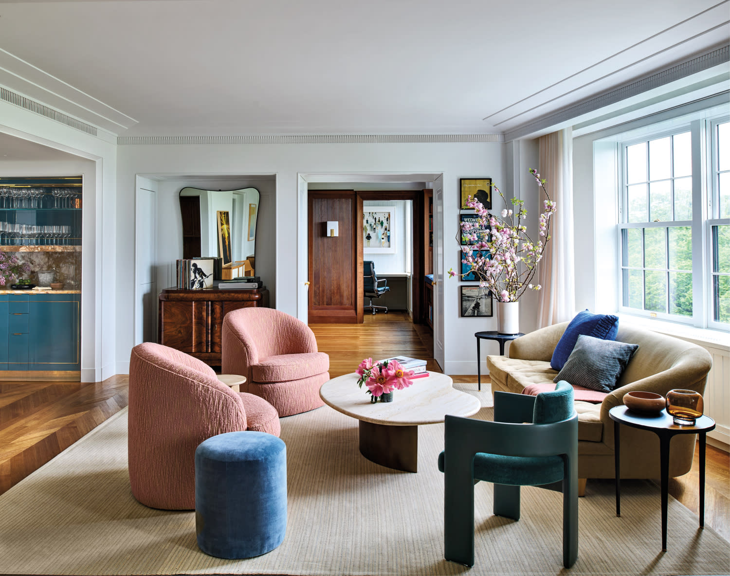A stylish living room with pastel tones features a beige sofa, pink armchairs, a round coffee table with pink flowers, and large windows, creating a cozy, elegant ambiance.