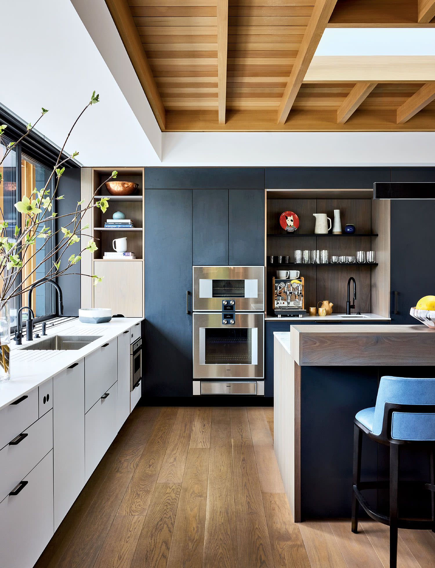 Modern kitchen with wooden ceiling beams, spacious white cabinets, dark backsplash, sleek appliances, a central island, and a blue stool. Bright and inviting.