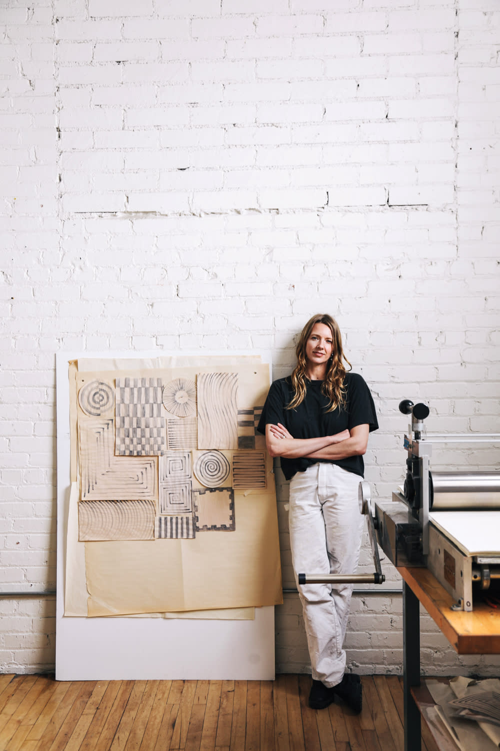 artist Cheryl Humphreys stands in front of a paper press and hand drawn sample works against a white brick wall