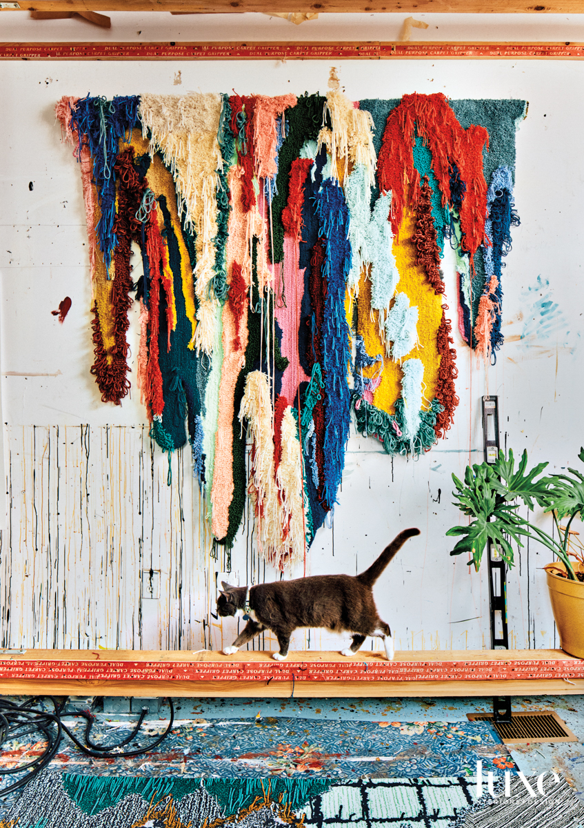 Multicolored, textural wall hanging above a bench with a strolling cat