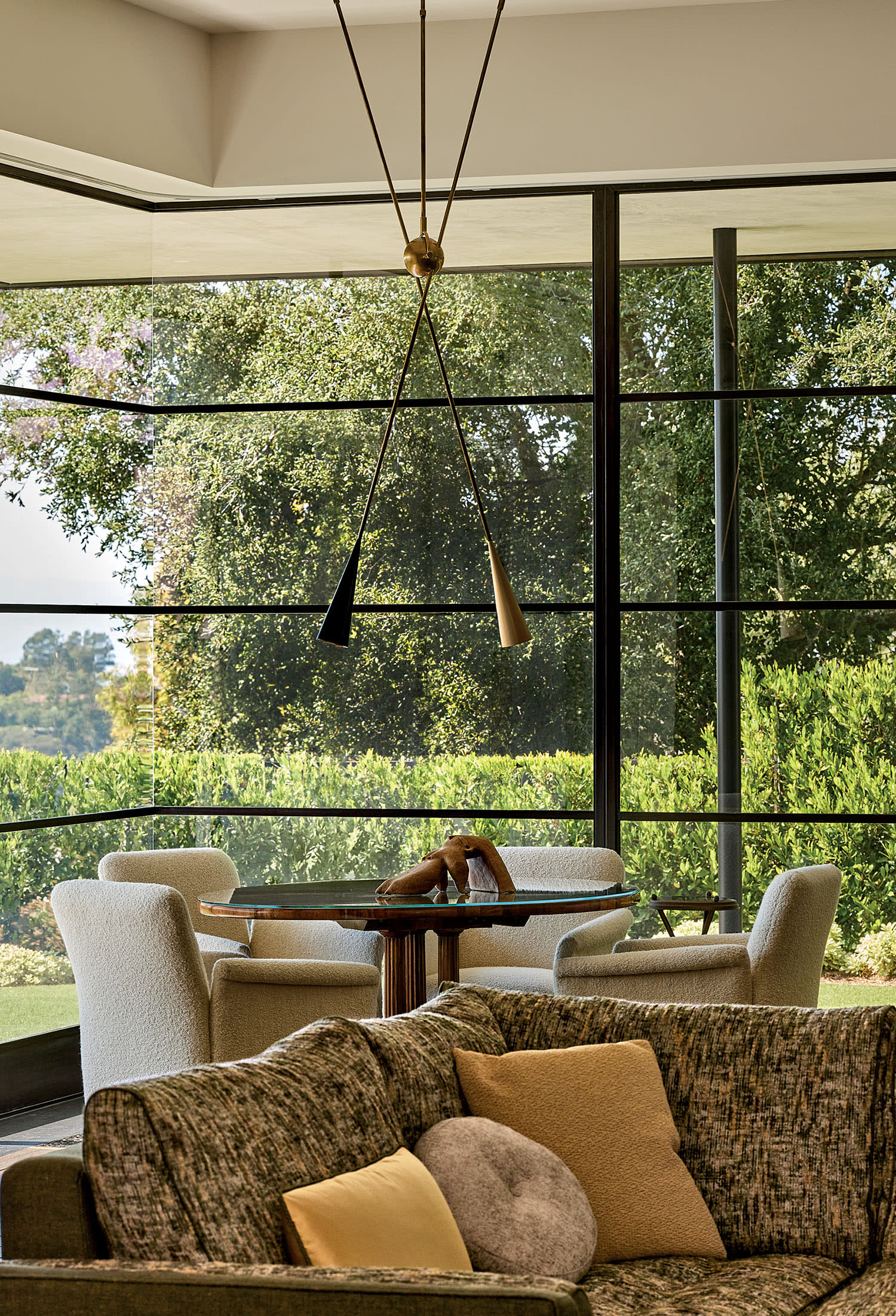 Modern living room with large windows revealing lush greenery. Features a cozy couch with cushions, round dining table, chairs, and stylish pendant light. Bright and serene ambiance.