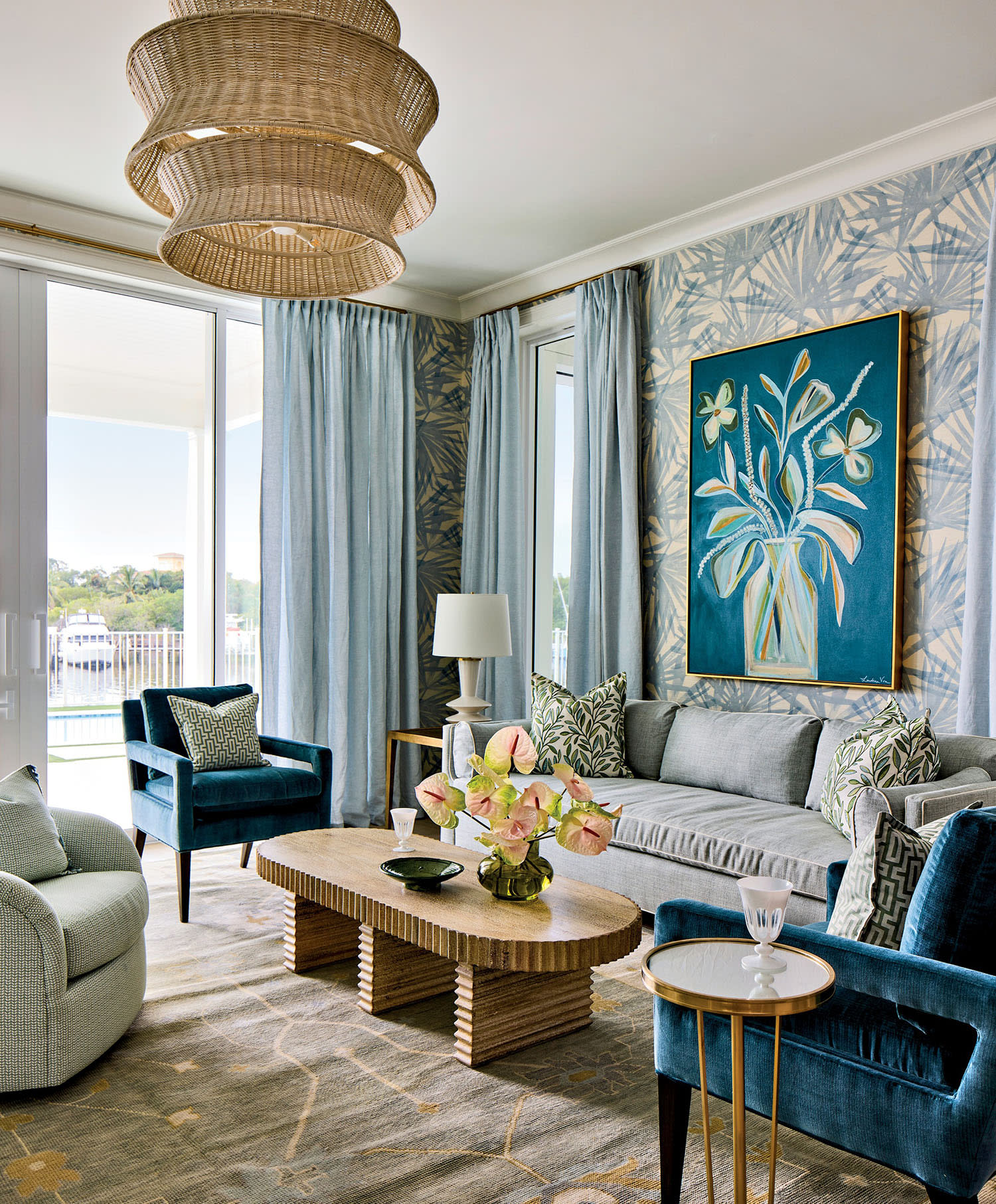 Elegant living room with blue velvet chairs, gray sofa, and modern art. Large rattan light fixture, floral decor, and patterned wallpaper accentuate calm ambiance.