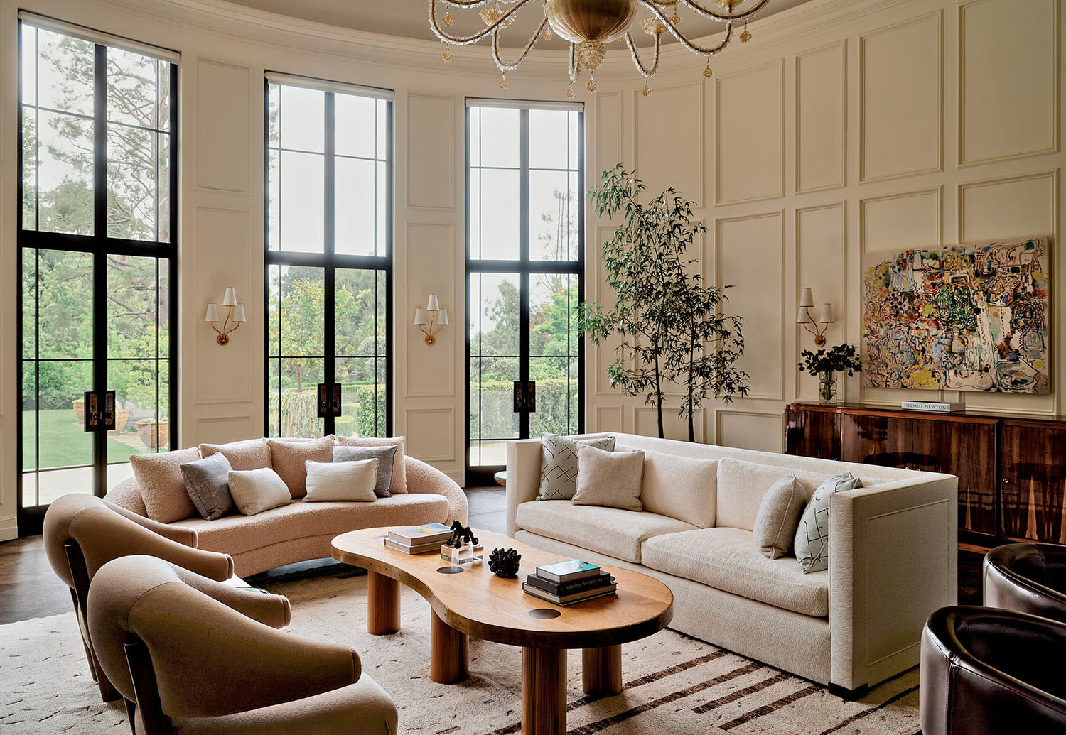 Spacious living room with large windows, beige sofas, a wooden coffee table, and a decorative chandelier. The space is bright, elegant, and inviting.