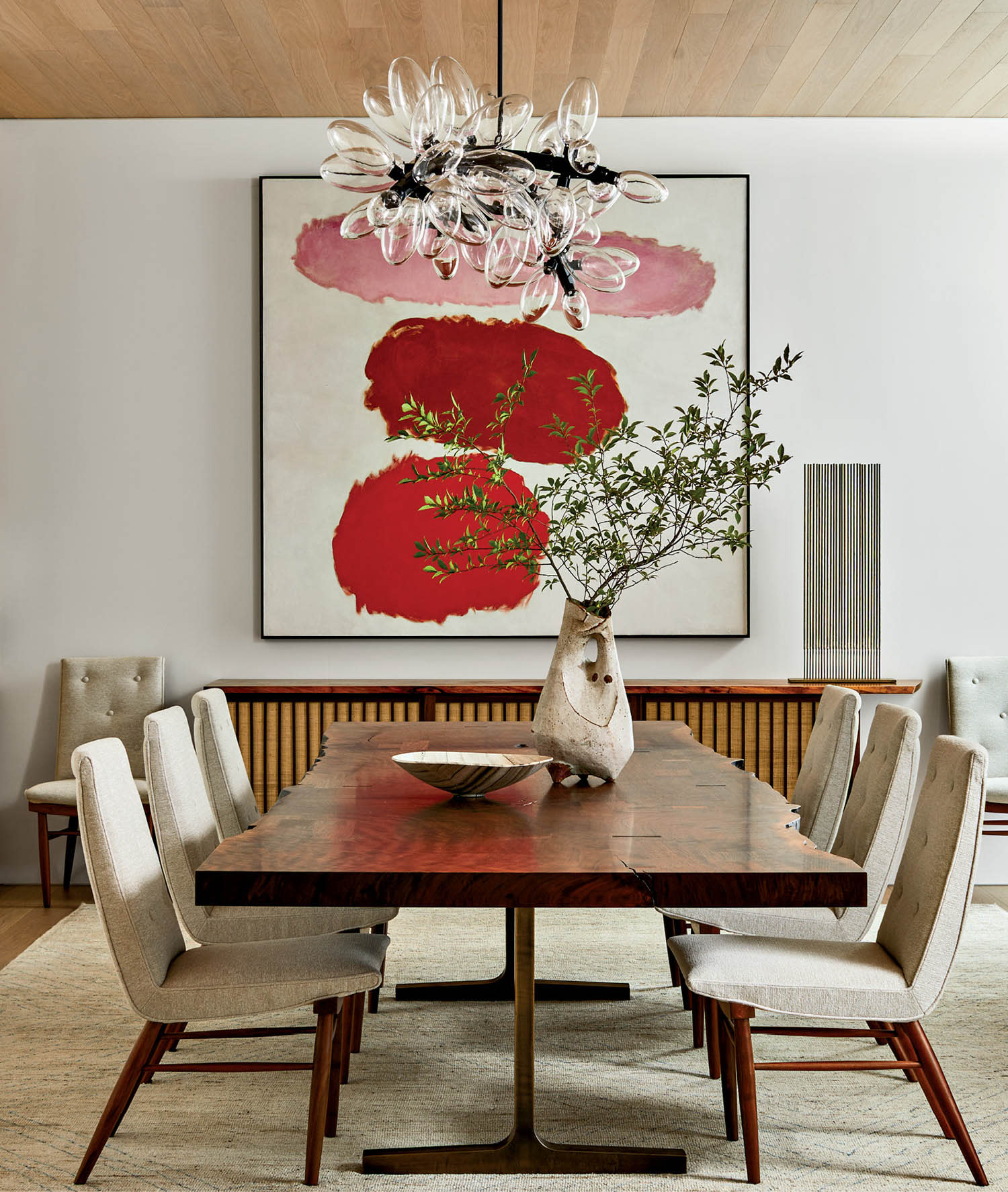 Sophisticated dining room with a wooden table, beige chairs, abstract red artwork, a modern glass chandelier, and a vase with green branches.