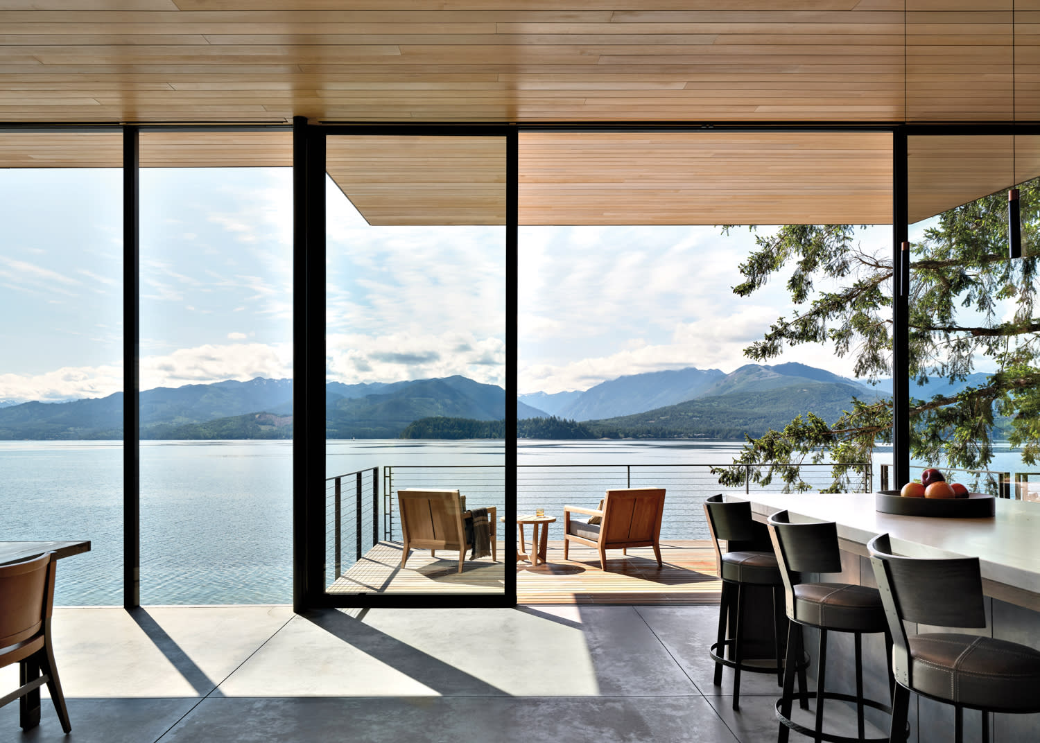 Modern interior with glass walls overlooking a serene lake and mountains. Patio features wooden chairs. The scene is calm and inviting.