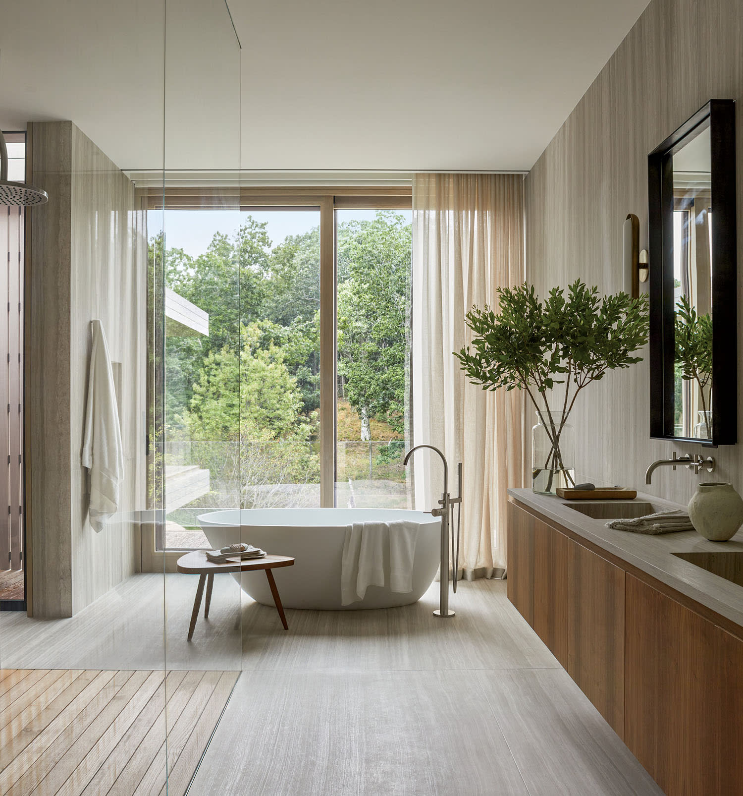 Elegant bathroom with a freestanding tub by large windows, sheer curtains, wooden accents, and neutral tones. A serene, spa-like atmosphere.