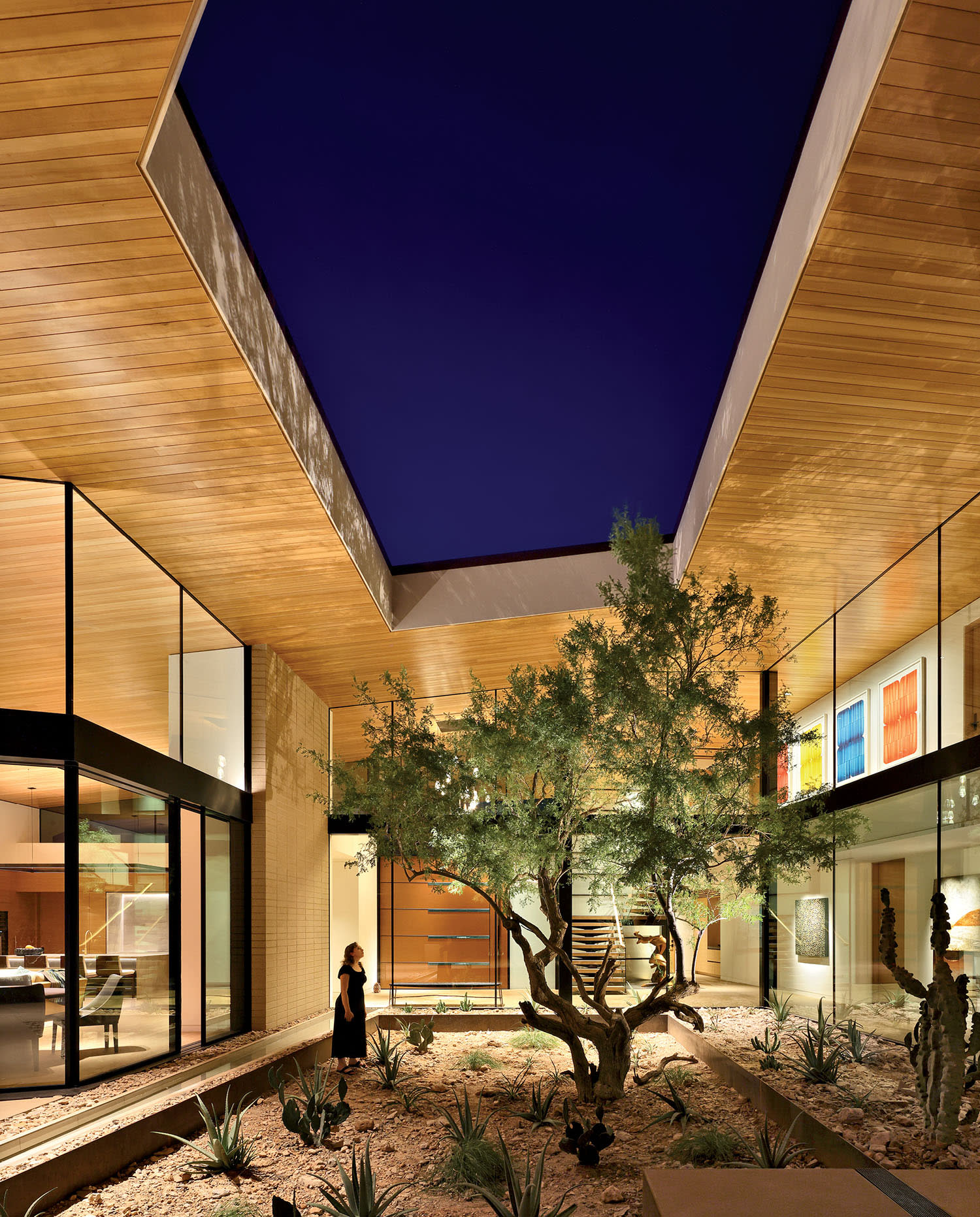 Modern courtyard with a central tree under a starry sky, surrounded by glass walls and warm wood ceilings. A person stands admiring the serene space.