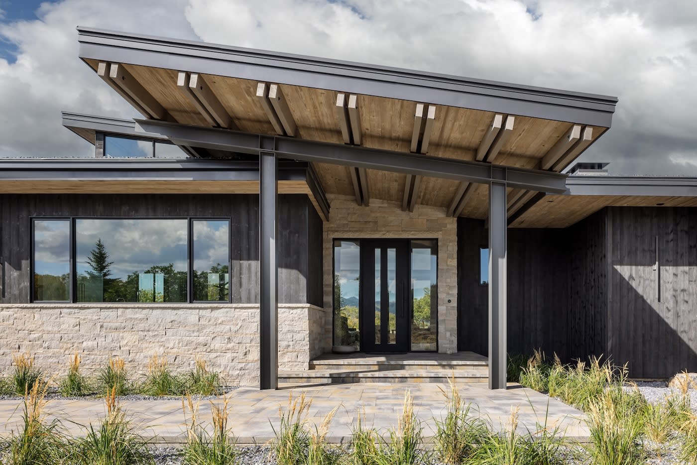 Modern house with a stone and dark wood facade, featuring a large overhanging roof. Glass door entrance, surrounded by tall grasses and cloudy sky.