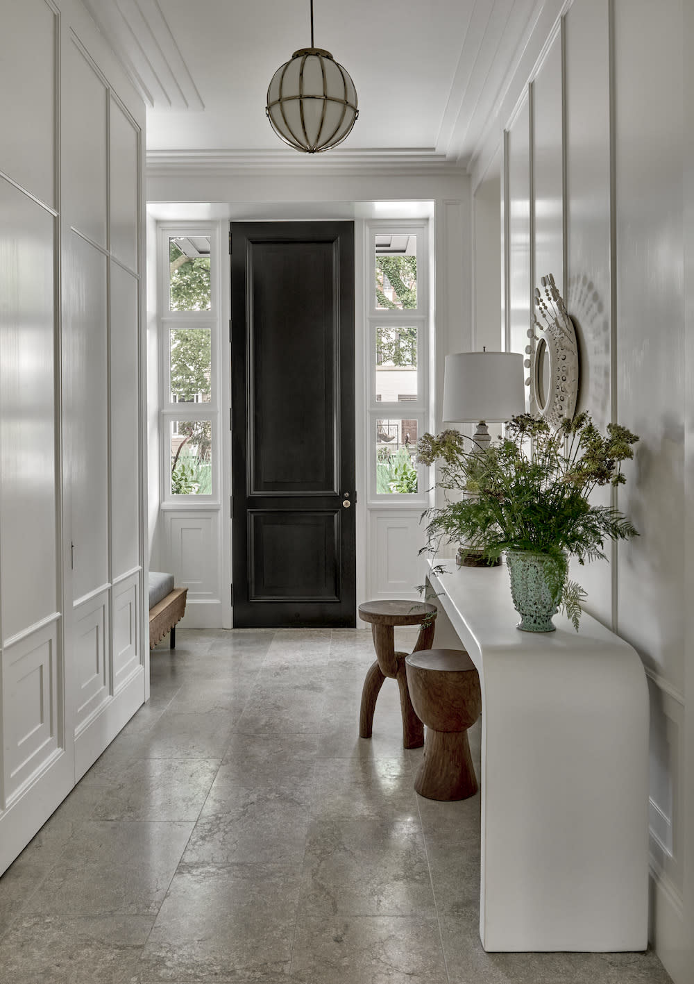 The combination of white paneled walls, stone tile flooring, a black door, and a singular pendant light creates a harmonious and inviting ambiance.