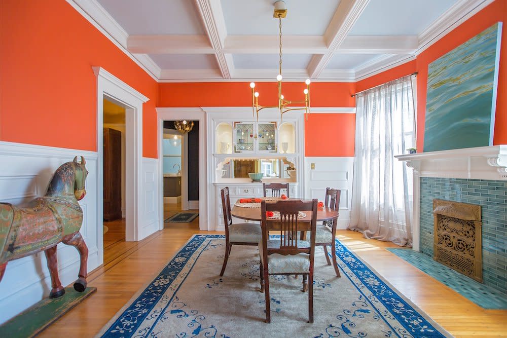 a vibrant dining room with warm orange walls providing a welcoming atmosphere. A sturdy wood table sits atop a blue area rug, anchoring the space with a pop of contrasting color. The white ceilings enhance the room's brightness, creating a visually pleasing and cozy dining environment.