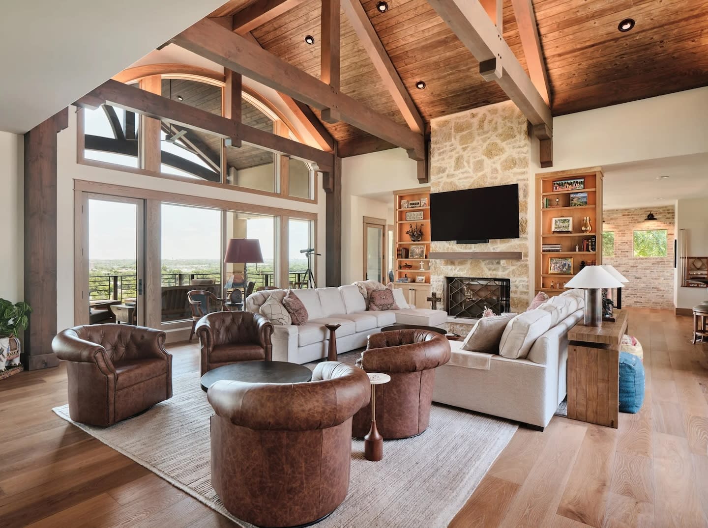 Spacious living room with rustic wood beams, stone fireplace, and large TV. Cozy seating includes a white sectional and leather chairs; natural light streams through large windows.