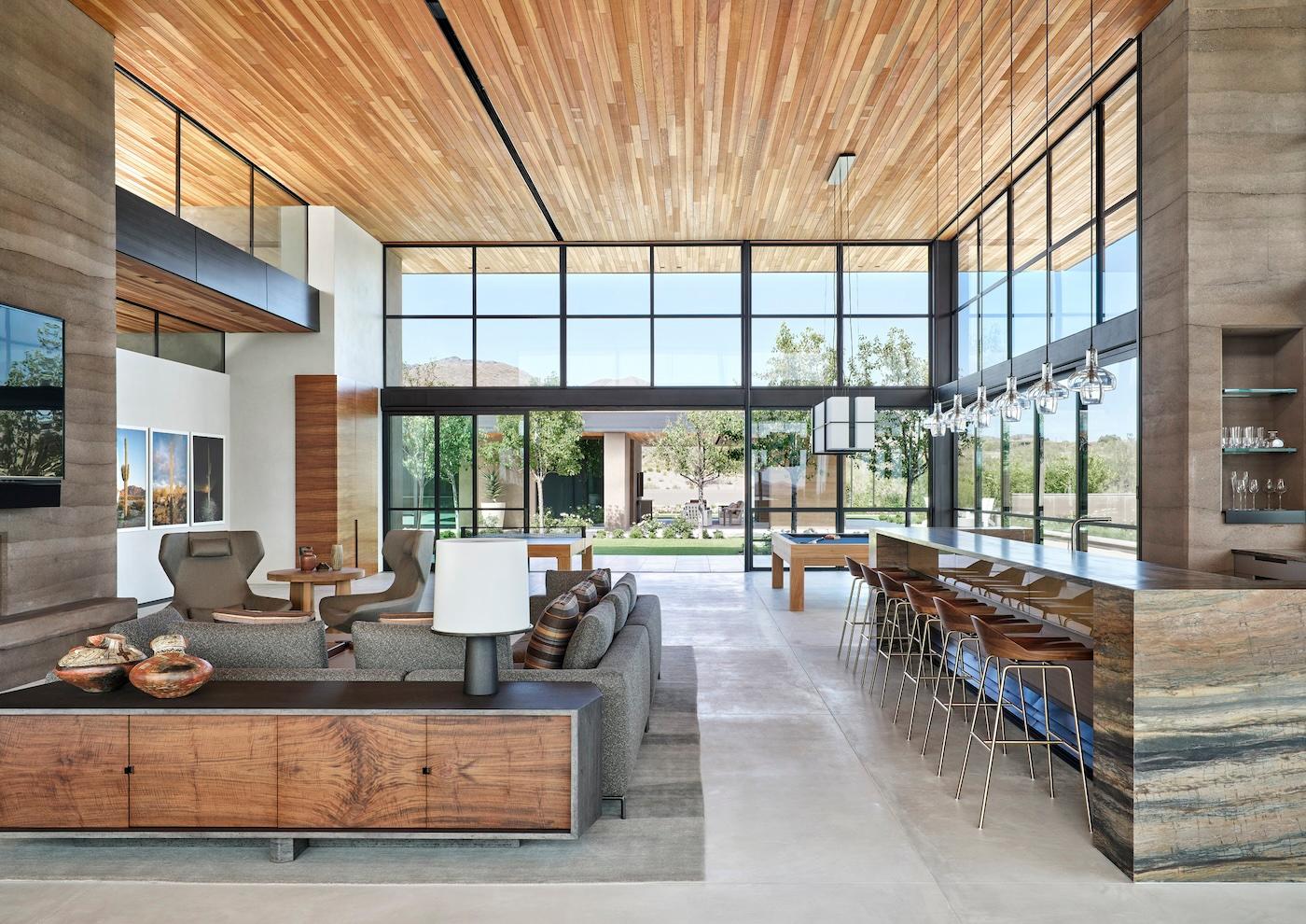 Modern open-concept living space with high wood ceilings, large windows, and neutral tones. Includes a sofa, chairs, bar area, and views of a garden.