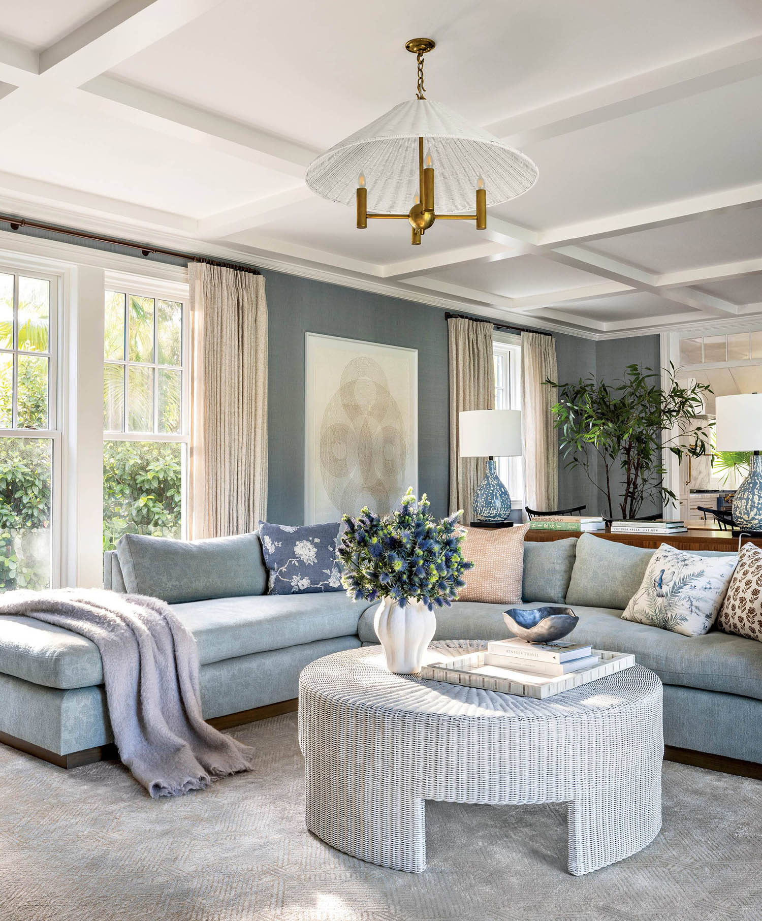 A curved couch decorated sits near a textured round coffee table in a room with large windows.