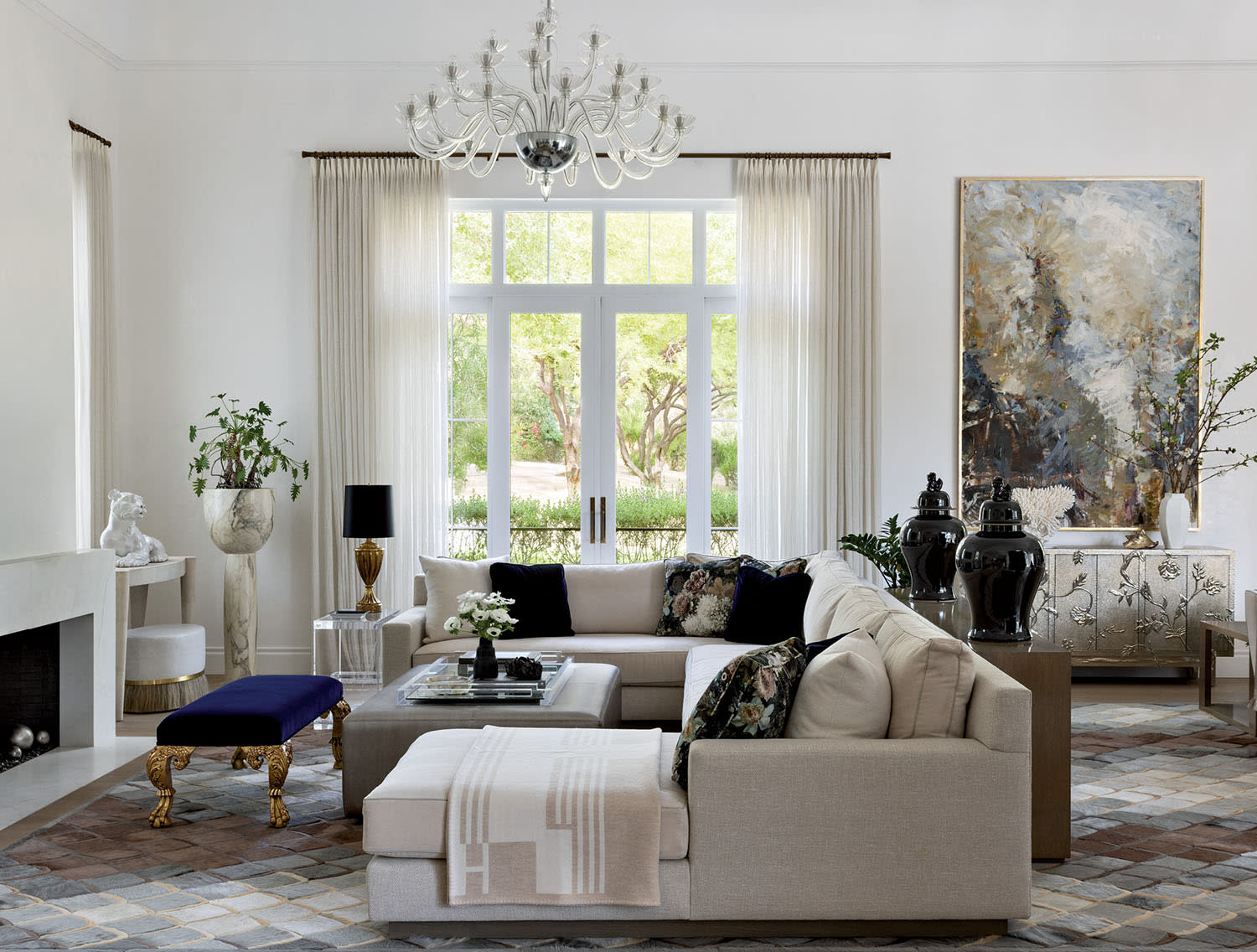 Elegant living room with a large beige sectional sofa, abstract art, and chandelier. Light from tall windows creates a serene, sophisticated ambiance.