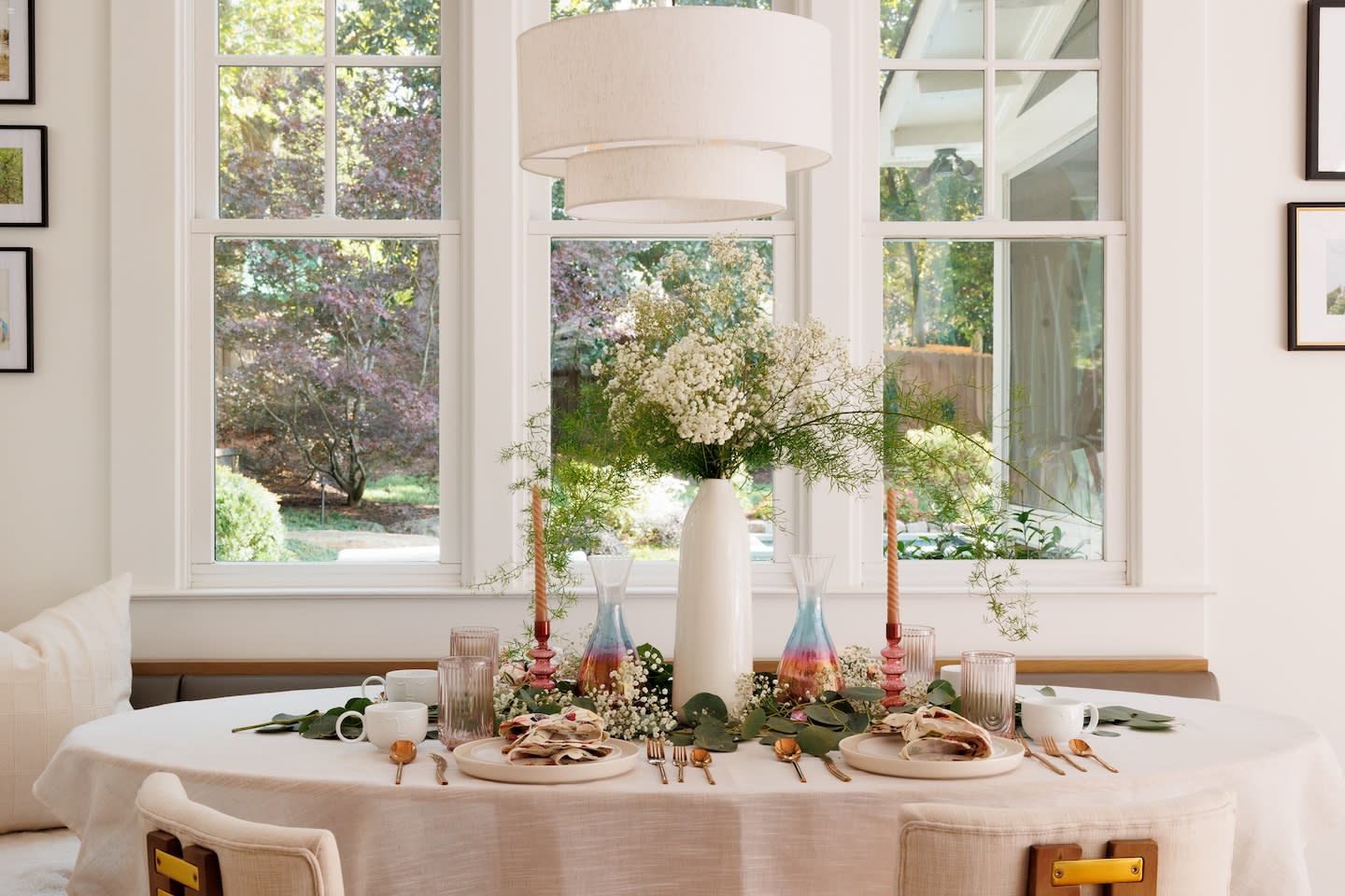 Elegant dining area with a table set for two, featuring white linens, colorful glassware, floral centerpiece, and greenery; sunlight streams through large windows.